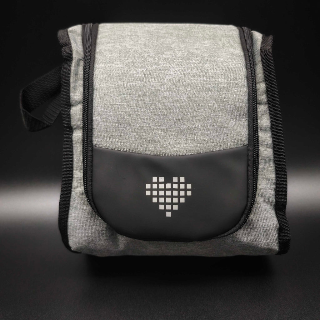 Eine stehende, graumelierte Kosmetiktasche mit schwarzem Reißverschluss und Tragegriff. Auf der Vorderseite befindet sich eine aufgesetzte, halbrunde, schwarze Tasche mit einem weißen, gepunkteten Herzsymbol in der Mitte. Der Hintergrund ist dunkelgrau bis schwarz.