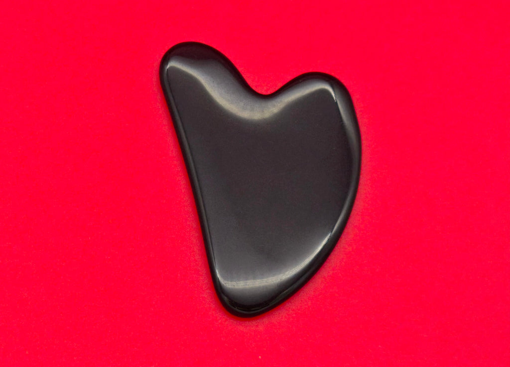 Ein glänzender, herzförmiger, schwarzer Gua-Sha-Massagestein liegt auf einem leuchtend roten Hintergrund.