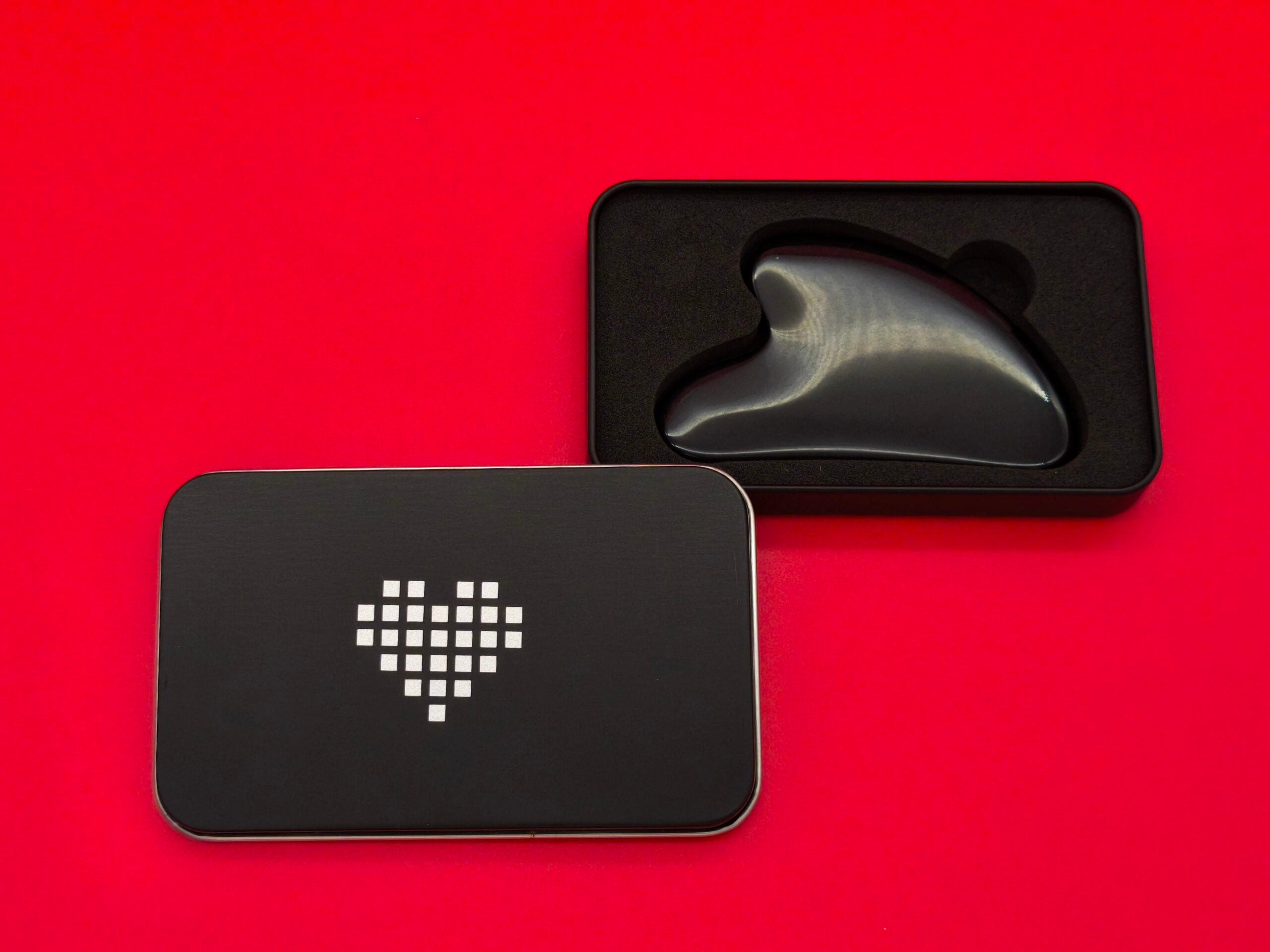 Ein schwarzer, herzförmiger Gua Sha Massagestein in einer schwarzen Metalldose mit abgerundeten Ecken. Der Deckel der Dose liegt daneben und zeigt ein weißes Pixelherz. Der Hintergrund ist leuchtend rot.